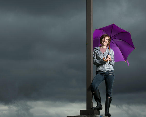 Professor Viviana is holding a purple umbrella and leaning against a pole while standing in front of cloudy skies.